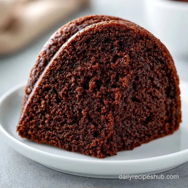 A slice of moist dark chocolate cake on a white plate, topped with a dollop of cream and a single fresh raspberry.